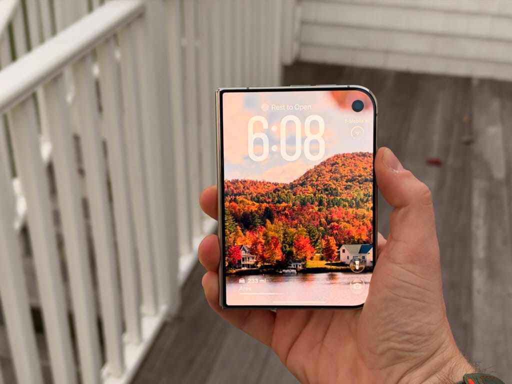A folded iPhone Duo held outdoors, showing its front lock screen and autumn wallpaper.